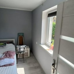 WOJT-KRAZ - Pomalowane na szaro ściany i biały sufit w pokoju, widoczne białe drzwi z klamką, okno z butelką na parapecie, łóżko z białą pościelą i stolik nocny, podłoga wyłożona panelami.