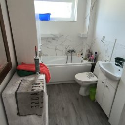 WOJT-KRAZ - Jasna łazienka z wanną obłożoną marmurową płytką, umywalką z szafką, toaletą z dekoracją kwiatową, szarą podłogą imitującą drewno i skrzynią z napisem jako elementem dekoracyjnym, widoczne ręczniki...