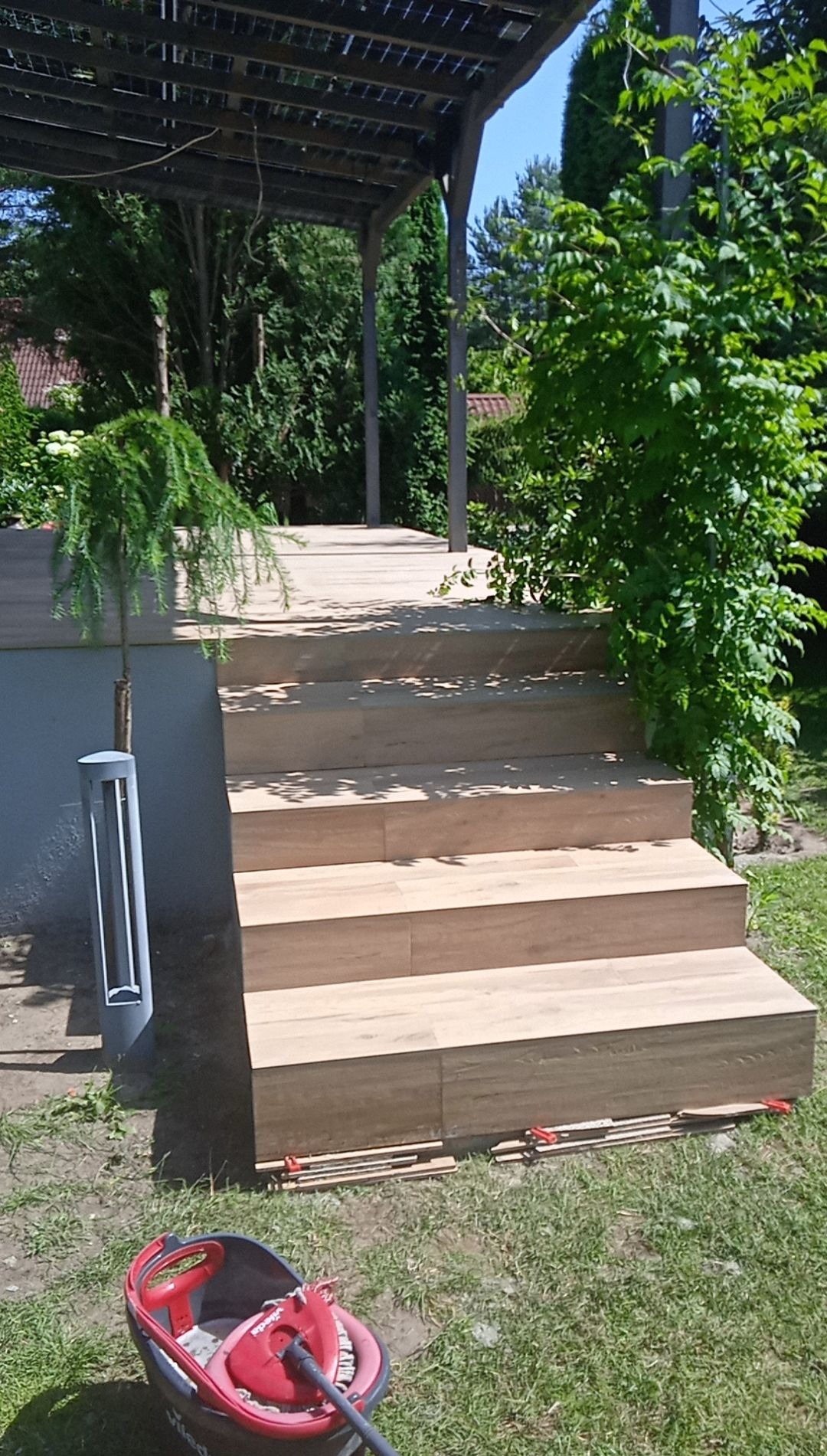 Zewnętrzne schody z płytek imitujących drewno prowadzące na taras z zadaszeniem, widoczne narzędzia i resztki materiałów po montażu.