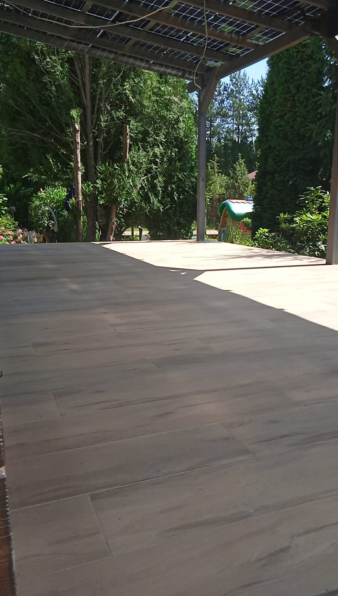 Zewnętrzna drewnopodobna podłoga tarasowa pod zadaszeniem z panelami, widok na zielony ogród z drzewami iglastymi i małym domkiem dla dzieci w tle.