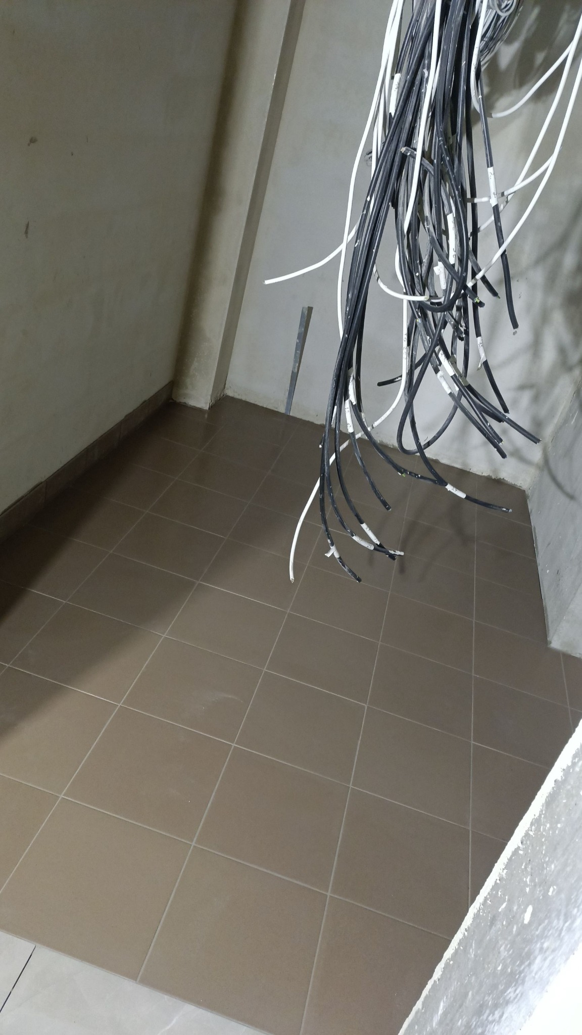 Łazienka w trakcie remontu: ułożona brązowa terakota na podłodze, wiszące kable elektryczne z oznaczeniami, surowe ściany w beżowym odcieniu.