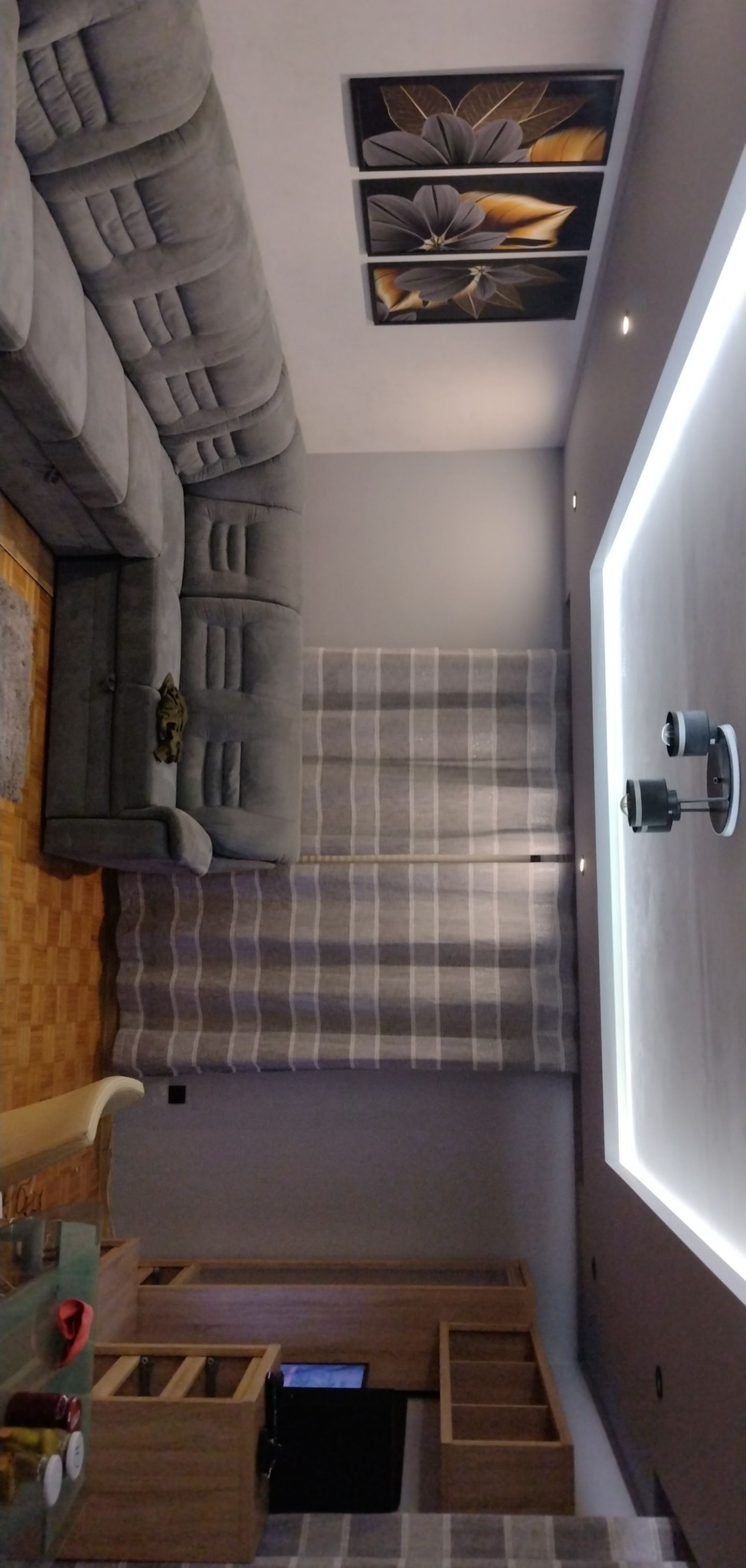 Nowoczesny salon z szarą, narożną sofą, panelami ściennymi z motywem kwiatowym, oświetleniem LED w suficie podwieszanym i drewnianymi meblami na tle szaro-białych zasłon.