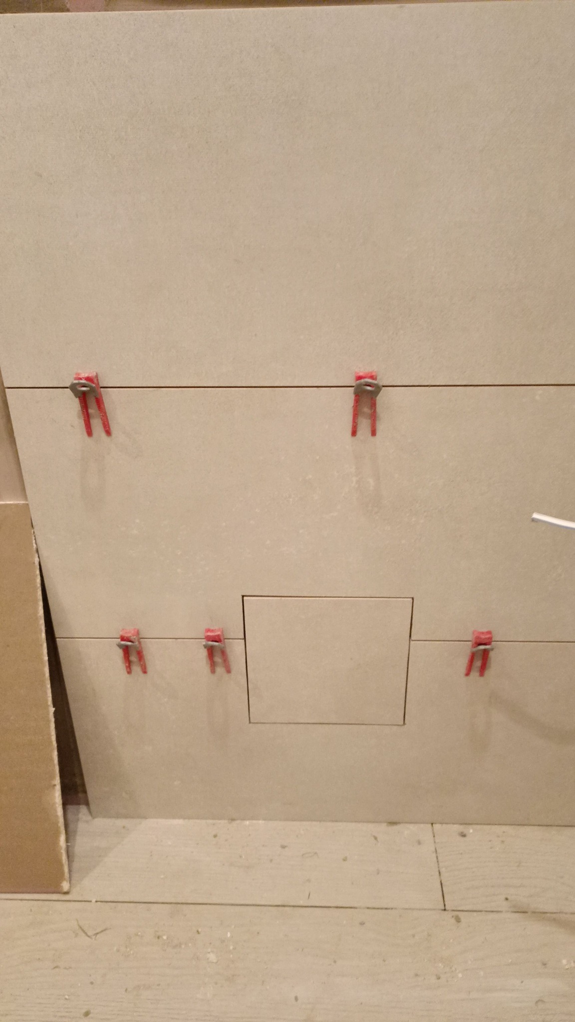 Układanie beżowych płytek z plastikowymi krzyżykami dystansowymi, widoczny otwór rewizyjny, podłoga z jasnych paneli.