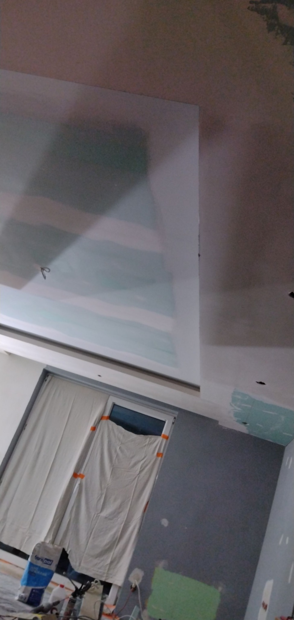 Sufit i ściany w trakcie szpachlowania, widoczne łączenia płyt kartonowo-gipsowych, okno zasłonięte folią ochronną, na podłodze worek z gładzią i narzędzia.