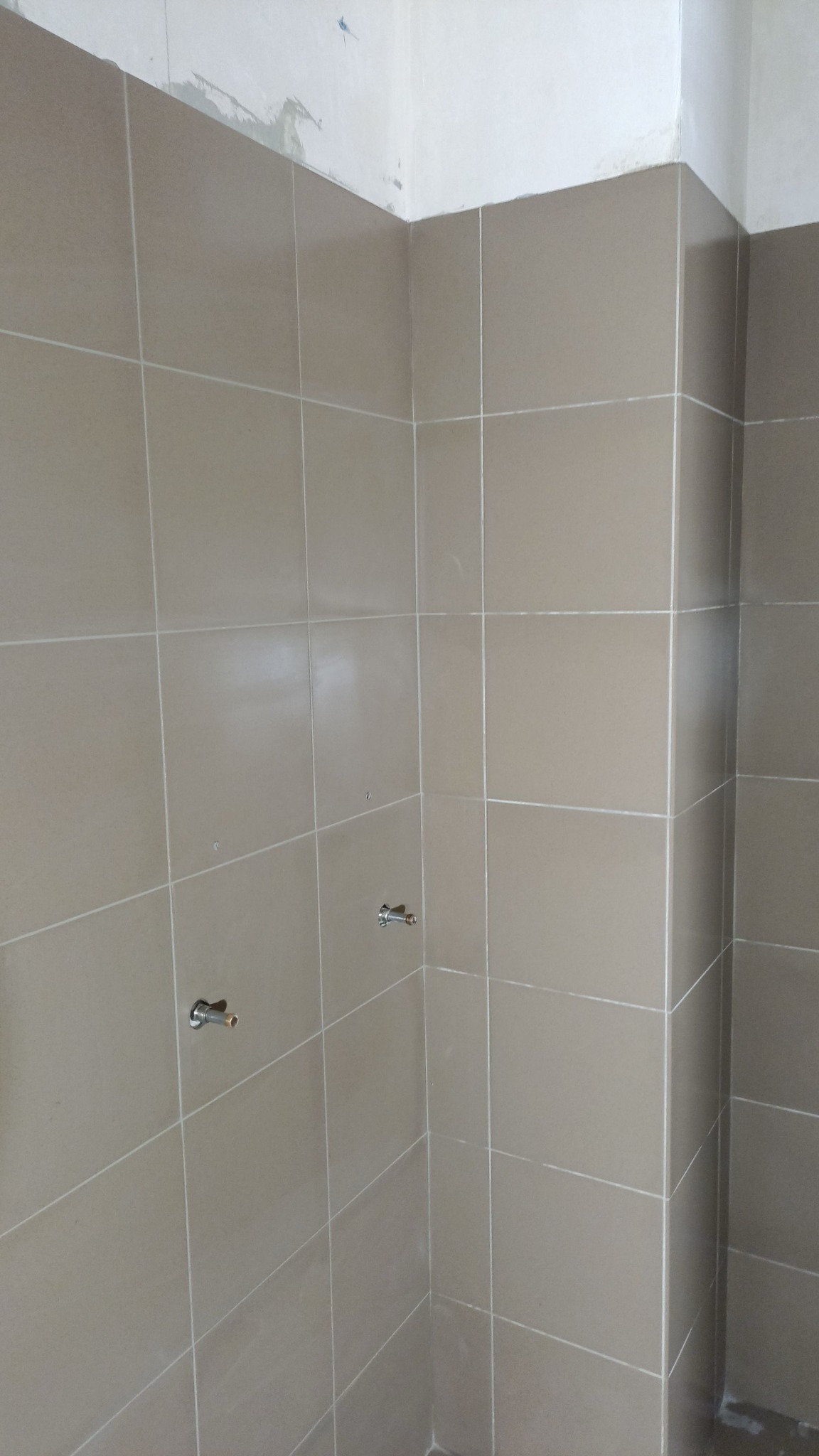 Ściana łazienki wyłożona beżowymi płytkami ceramicznymi, widoczne dwa wystające zawory wodne, ściana w trakcie prac wykończeniowych, góra ściany jeszcze nie wykończona.