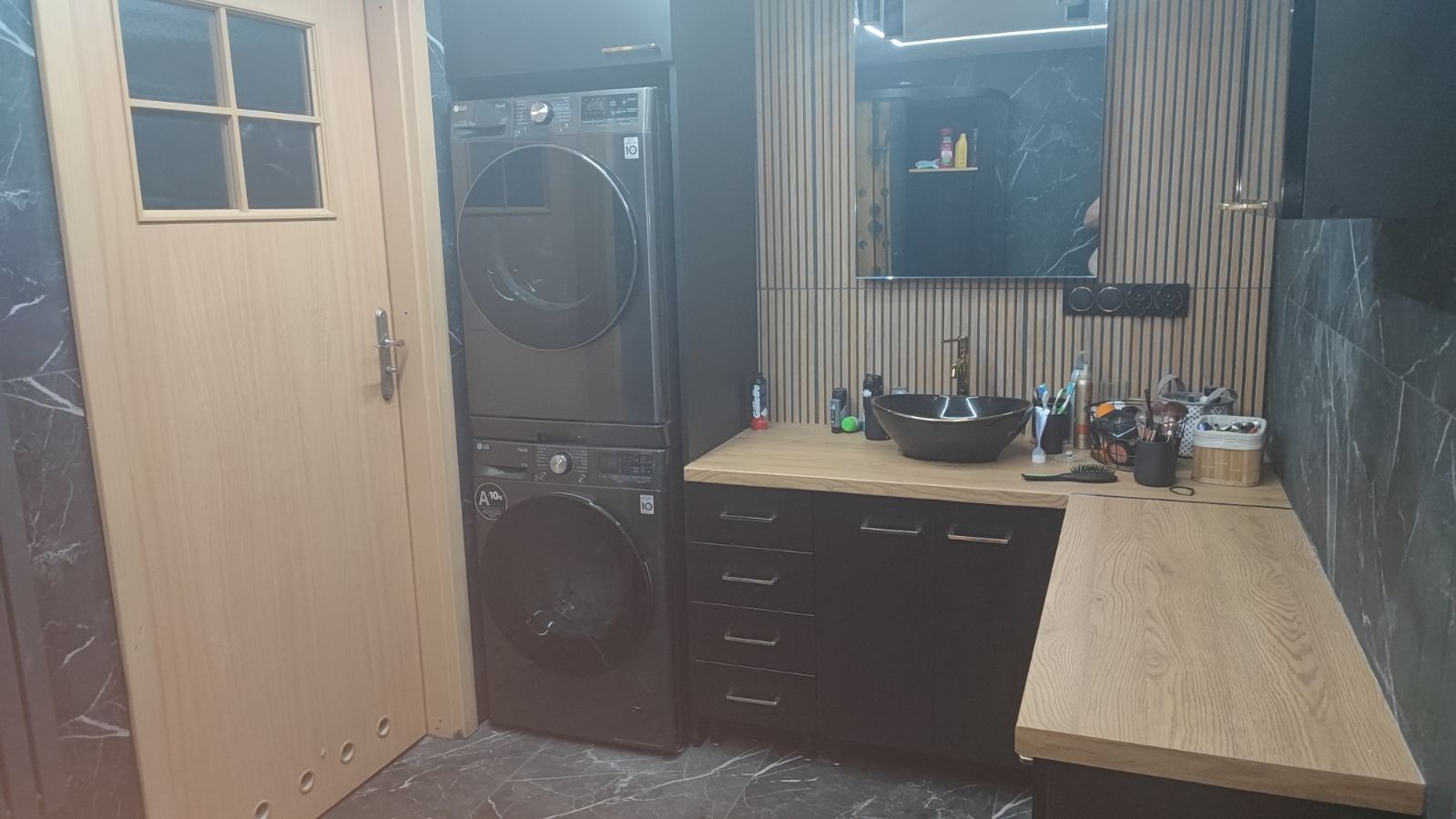 Nowoczesna łazienka z pralko-suszarką LG w kolorze grafitowym, umywalką nablatową w kształcie misy i drewnianymi lamelami dekoracyjnymi na ścianie za lustrem.