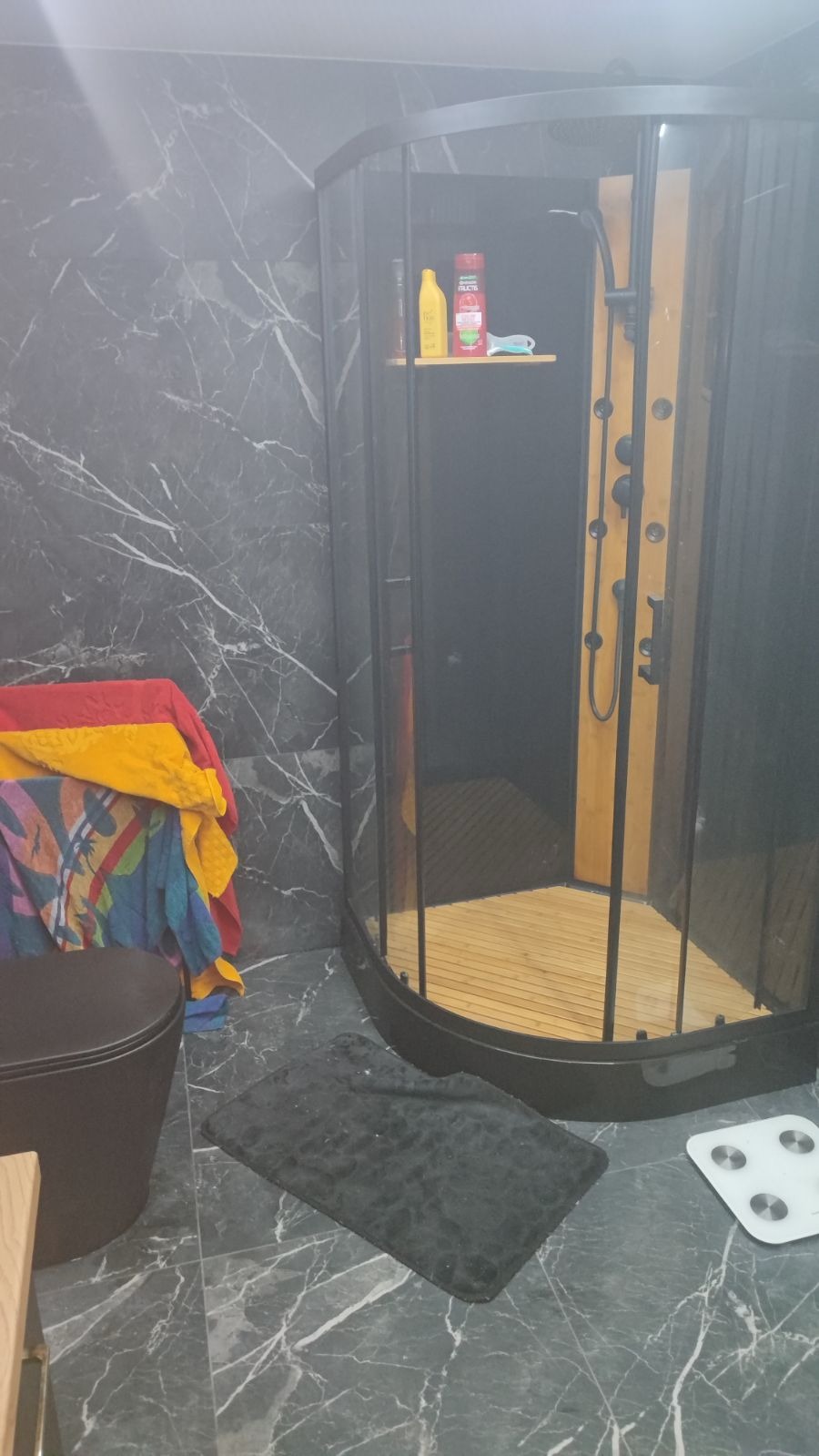 Nowoczesna kabina prysznicowa z hydromasażem, drewnianym panelem i czarną armaturą, widoczna na tle marmurowej ściany i podłogi, obok toaleta w kolorze czarnym i ręczniki.