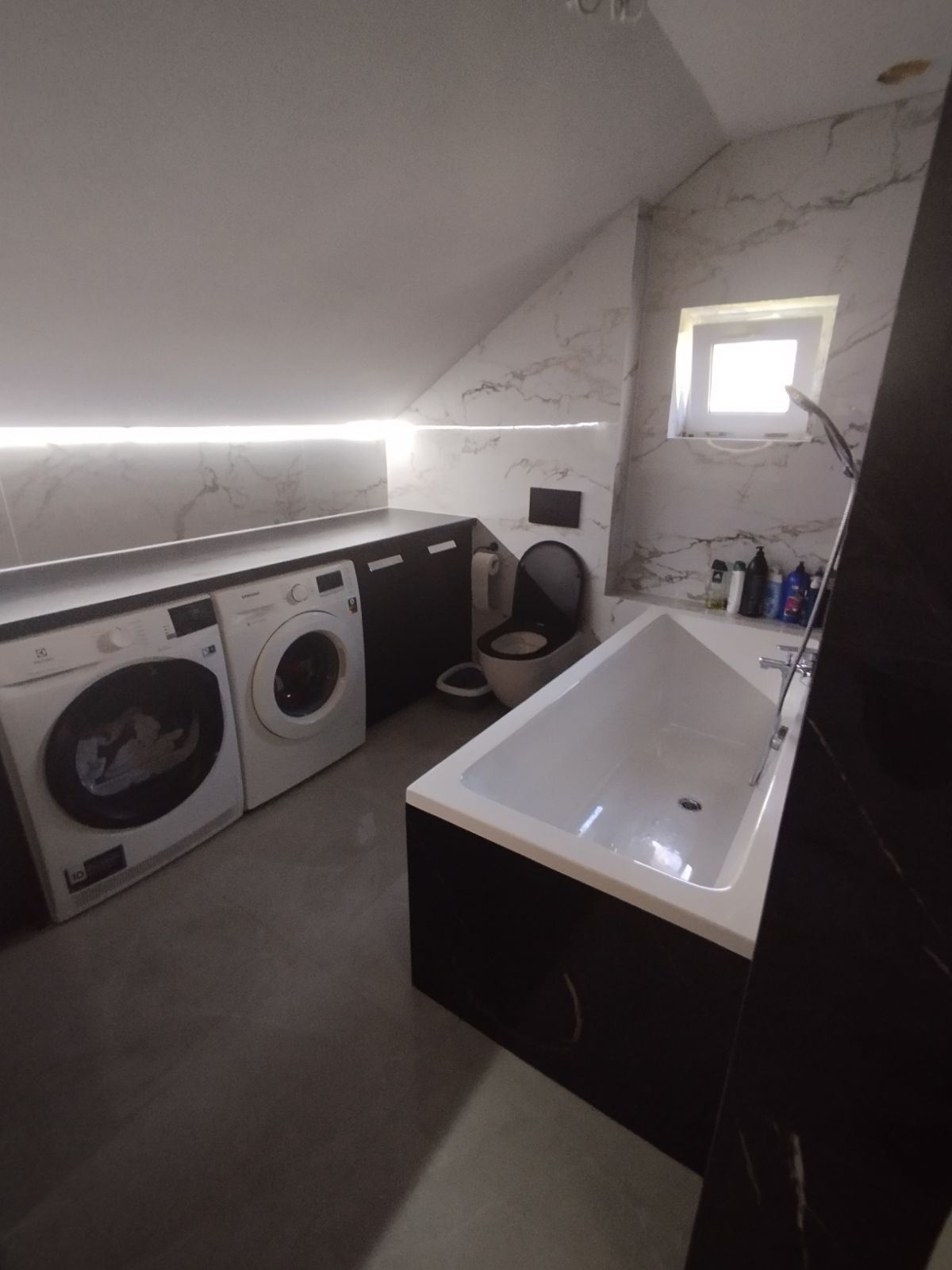 Nowoczesna łazienka na poddaszu z pralką, suszarką, wanną obudowaną czarną płytą i toaletą w kolorze czerni, wykończona marmurową płytą.