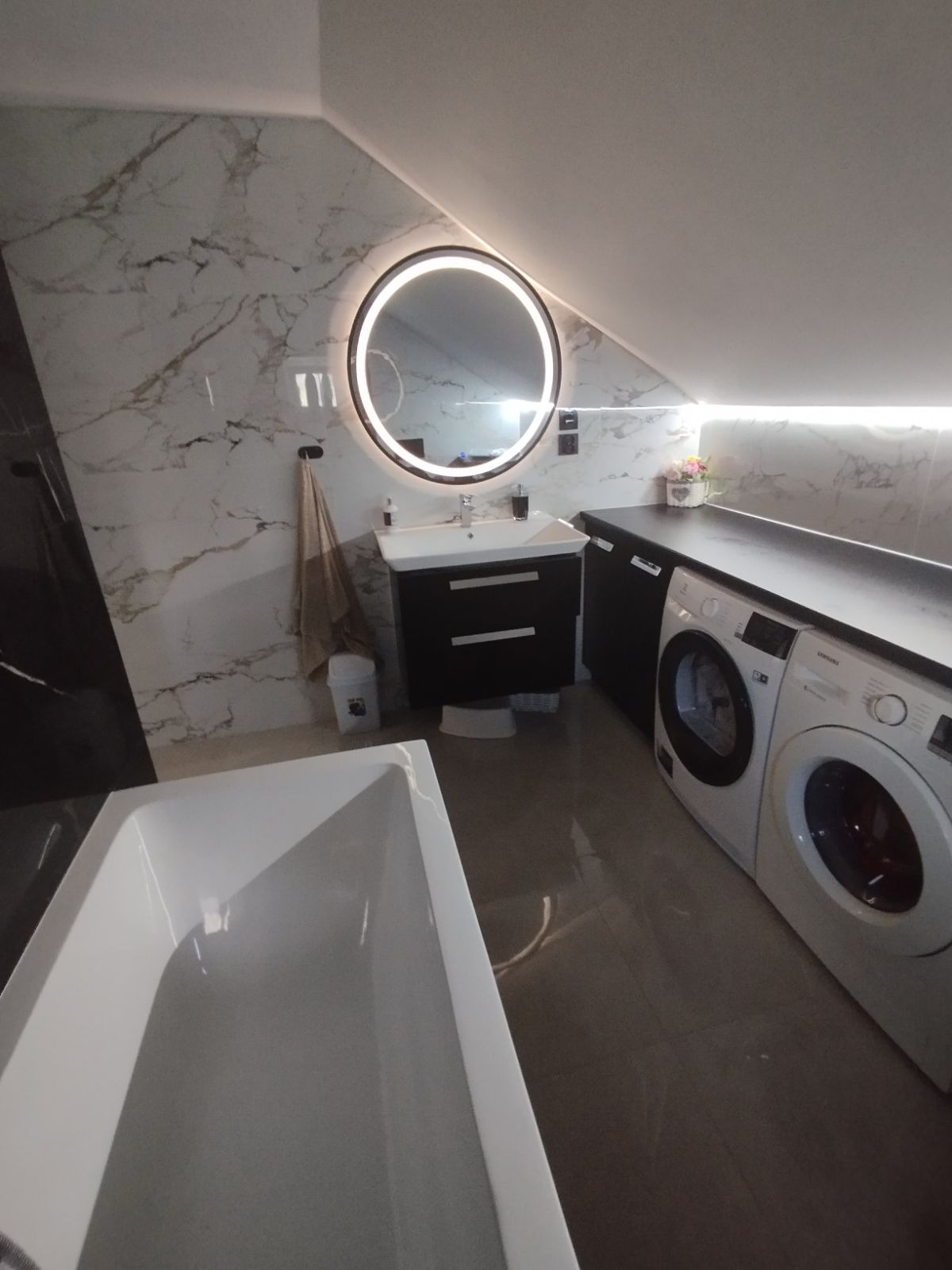 Nowoczesna łazienka na poddaszu z wanną, okrągłym lustrem z podświetleniem LED, umywalką z szafką i pralkami.