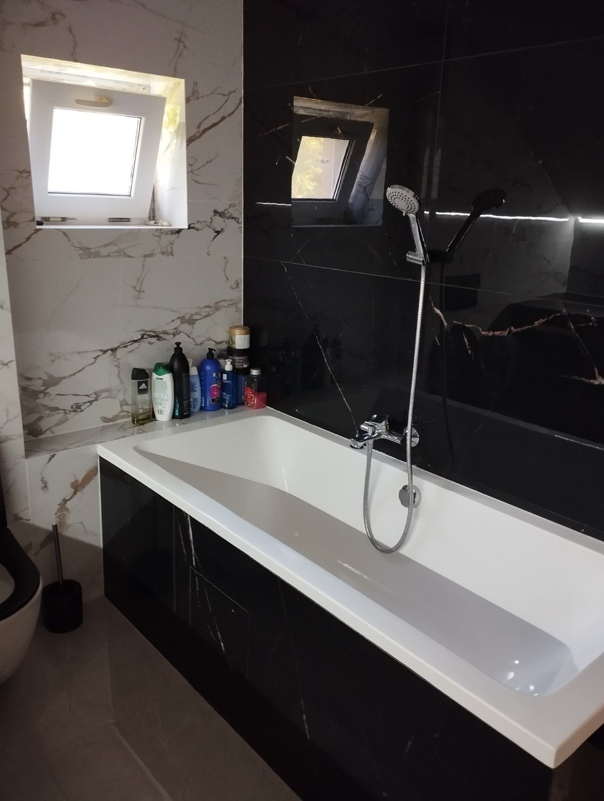 Biała wanna obudowana czarnymi płytami imitującymi marmur, chromowana bateria z prysznicem, biało-szare marmurowe płytki na ścianie, okno w górnym rogu, kosmetyki na półce.