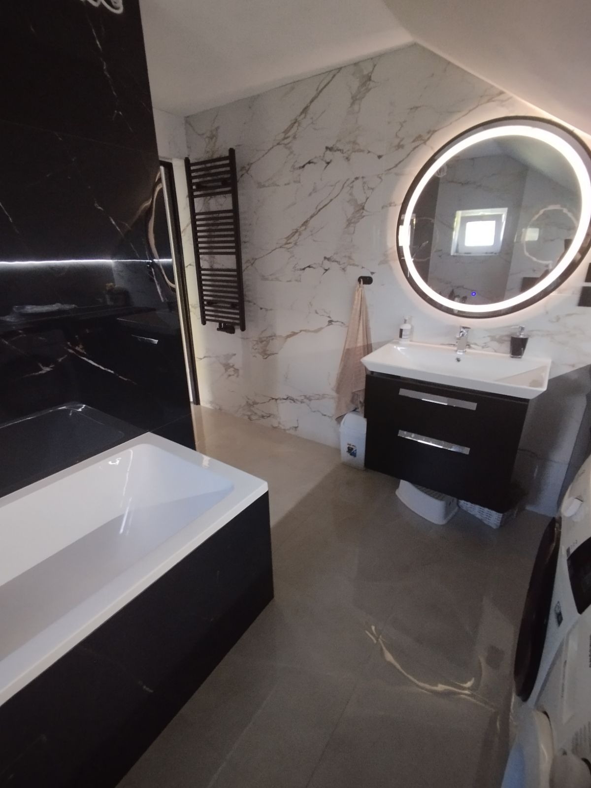 Nowoczesna łazienka w czarno-białej tonacji z marmurowymi płytkami, podświetlanym okrągłym lustrem, grzejnikiem drabinkowym i wolnostojącą wanną z czarną obudową.