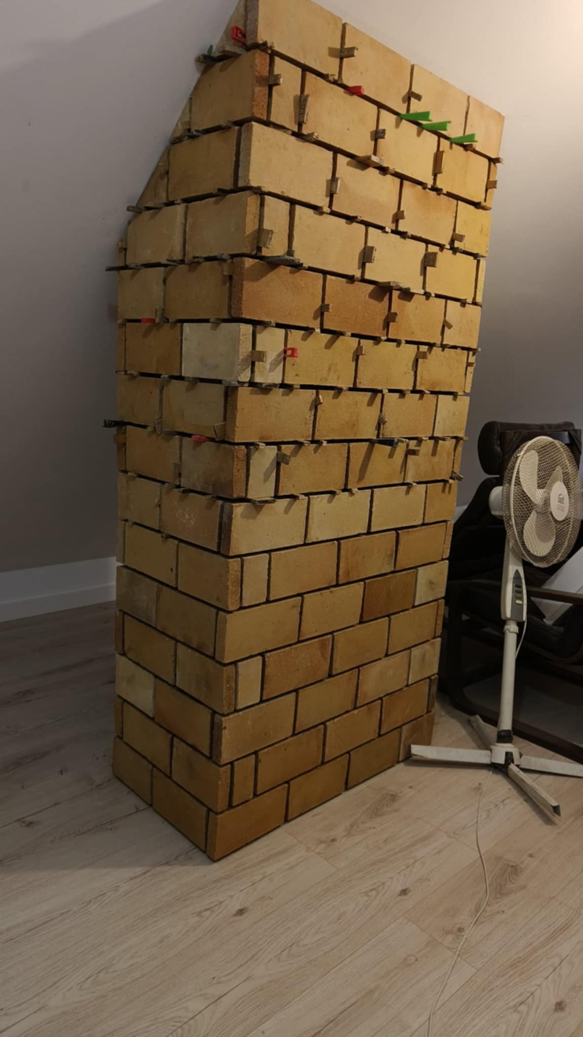 Wznoszenie konstrukcji z cegieł na poddaszu, z użyciem krzyżyków dystansowych, widoczny wentylator i skośny sufit.