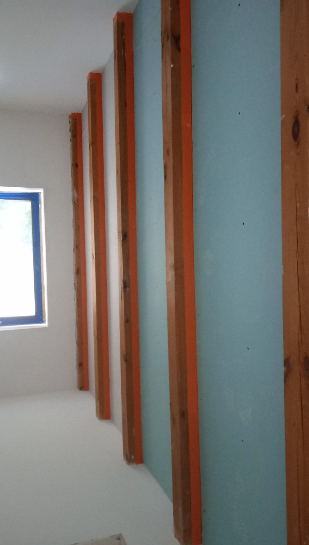 Ściana z zamontowaną niebieską płytą kartonowo-gipsową na drewnianym ruszcie z pomarańczowymi elementami mocującymi, widok z boku na jasne pomieszczenie z oknem.