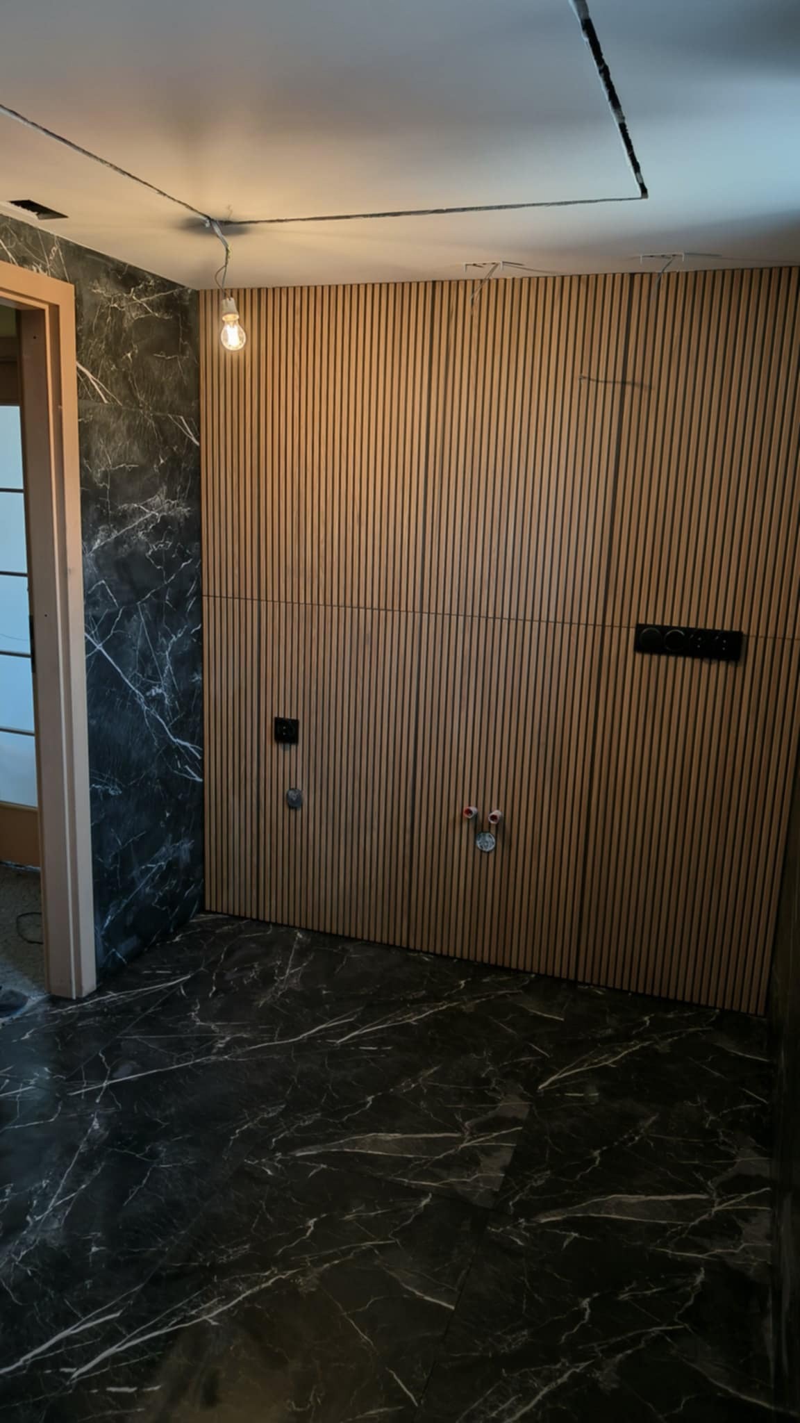 Nowoczesna łazienka w trakcie wykończenia: ściana z drewnianych paneli pionowych, czarna marmurowa podłoga, widoczne instalacje wodne i elektryczne, surowa żarówka na kablu.