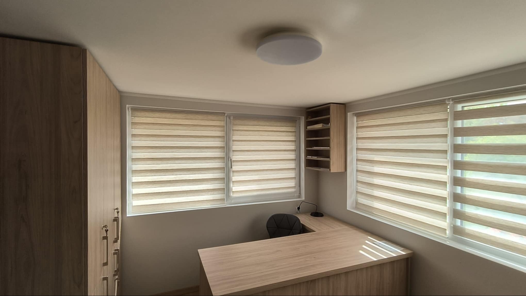 Jasne biuro z trzema oknami przysłoniętymi roletami dzień-noc, drewniane biurko narożne, szafka wisząca i wysoka zabudowa meblowa przy ścianie.