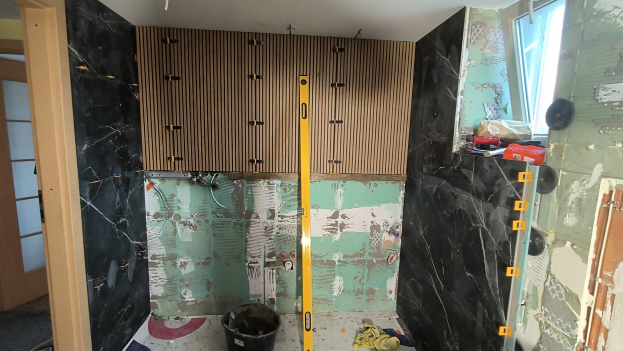 Remont łazienki w trakcie: montaż paneli ściennych z drewnianych listew oraz płytek imitujących czarny marmur, widoczny poziomica i narzędzia.