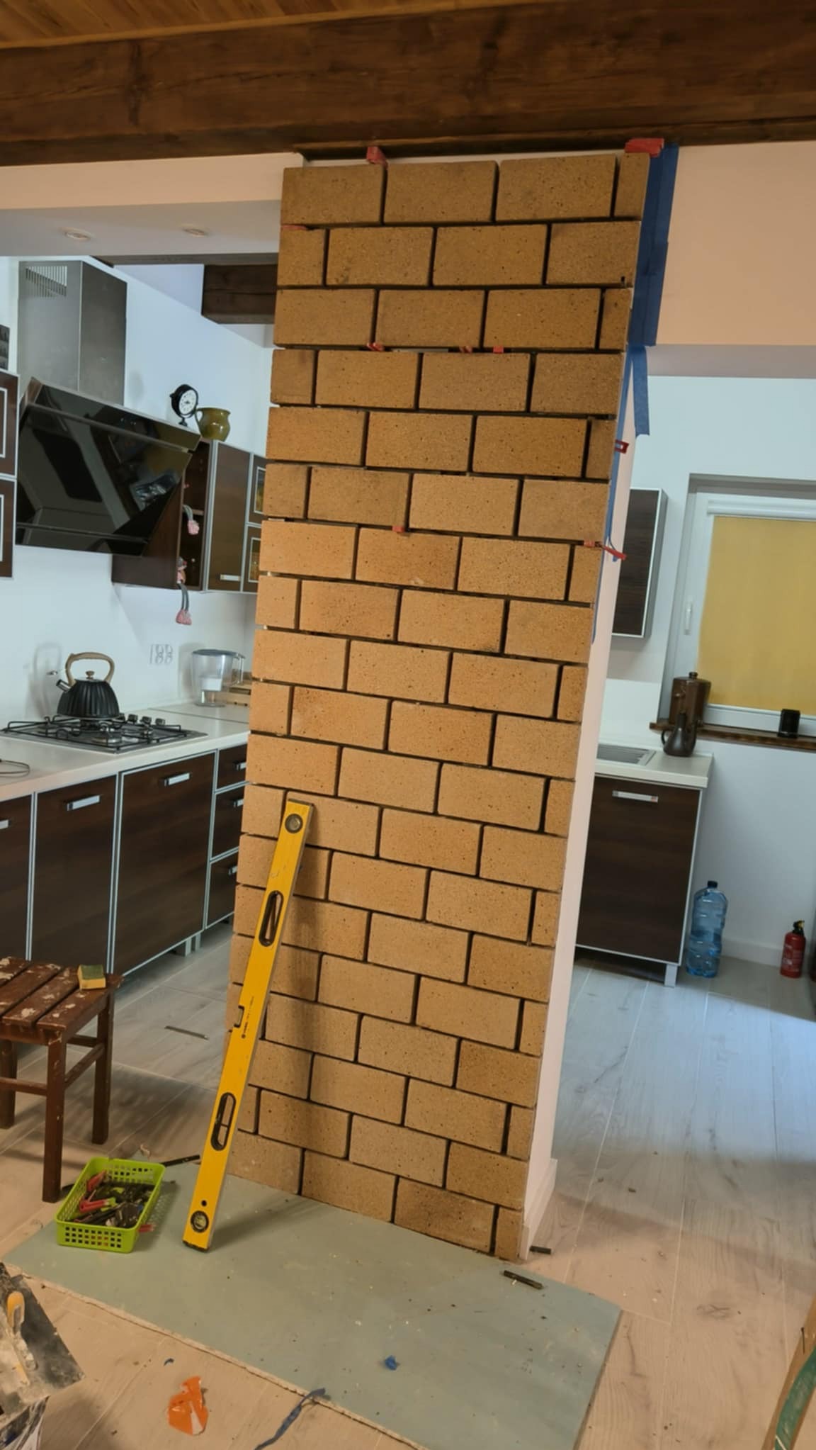 Wznoszenie ścianki działowej z cegły w kuchni, poziomica przyłożona do ściany, widoczne plastikowe krzyżyki dystansowe między cegłami, fragment zabudowy kuchennej w tle.