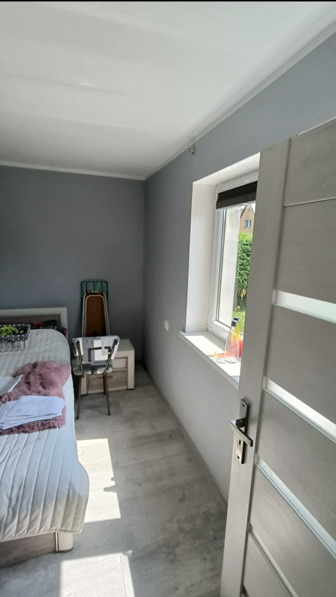 Pomalowane na szaro ściany i biały sufit w pokoju, widoczne białe drzwi z klamką, okno z butelką na parapecie, łóżko z białą pościelą i stolik nocny, podłoga wyłożona panelami.