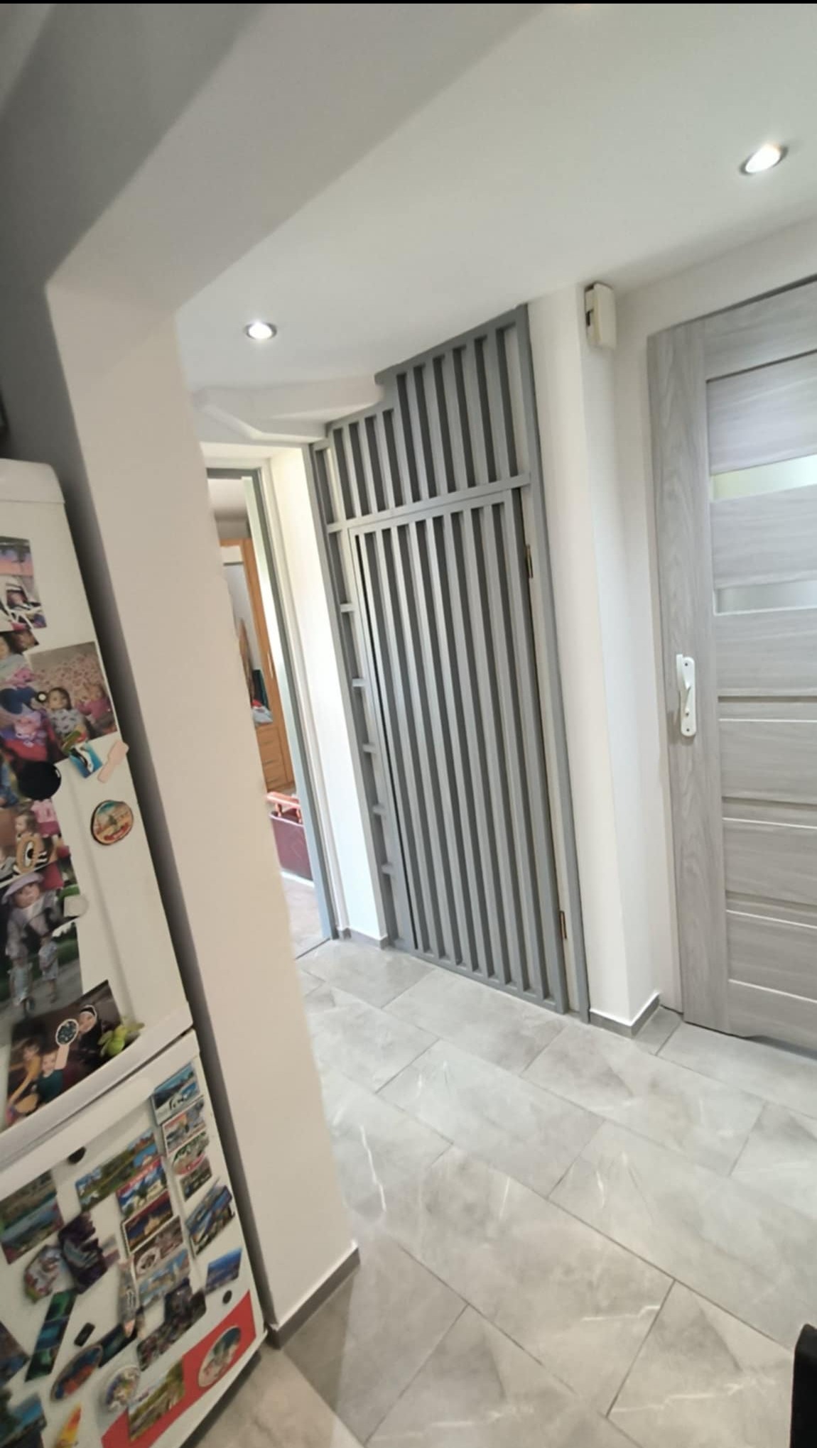 Szara, nowoczesna łazienka z płytkami imitującymi marmur, widoczna ażurowa ścianka działowa i drzwi w podobnej tonacji kolorystycznej, lodówka z magnesami w tle.