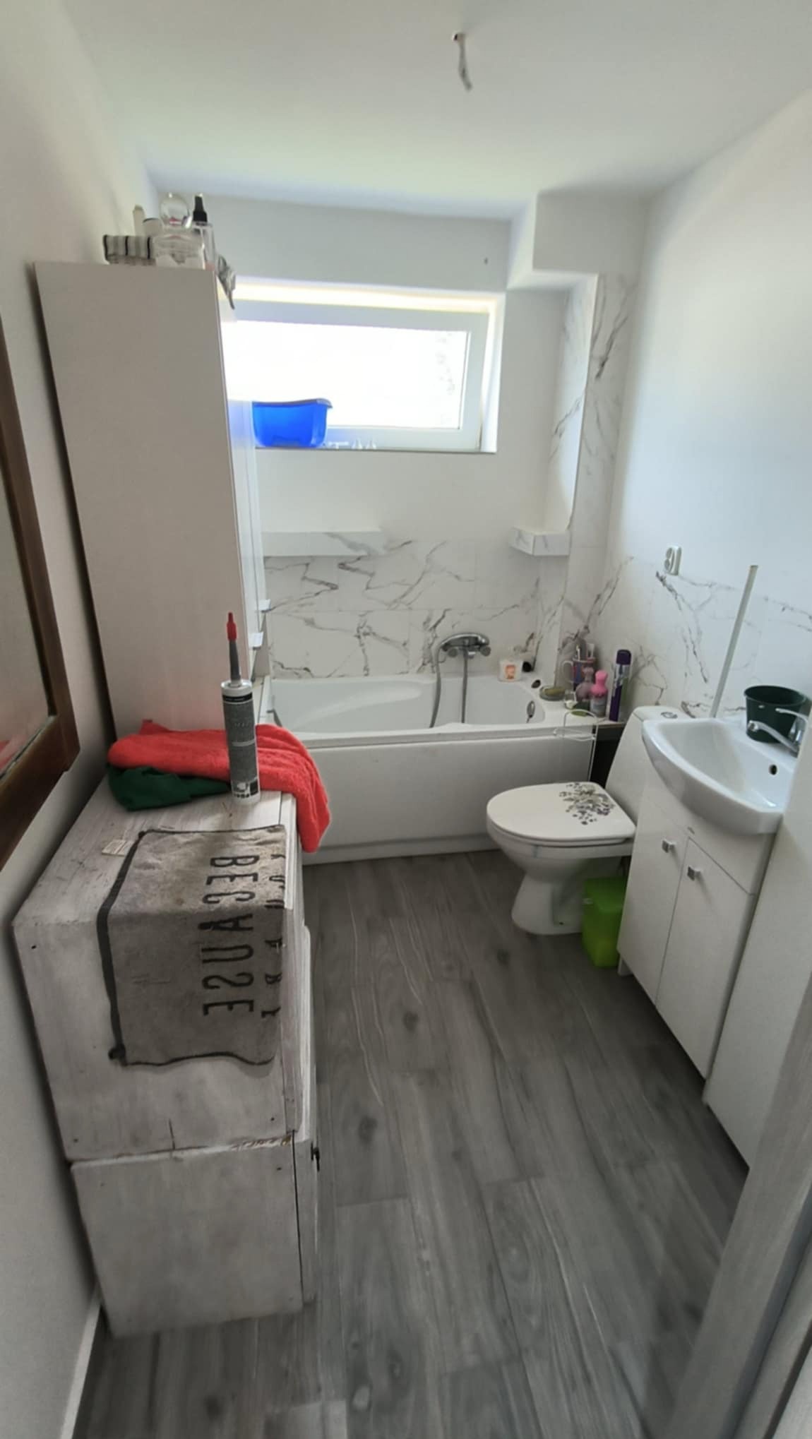 Jasna łazienka z wanną obłożoną marmurową płytką, umywalką z szafką, toaletą z dekoracją kwiatową, szarą podłogą imitującą drewno i skrzynią z napisem jako elementem dekoracyjnym, widoczne ręczniki...