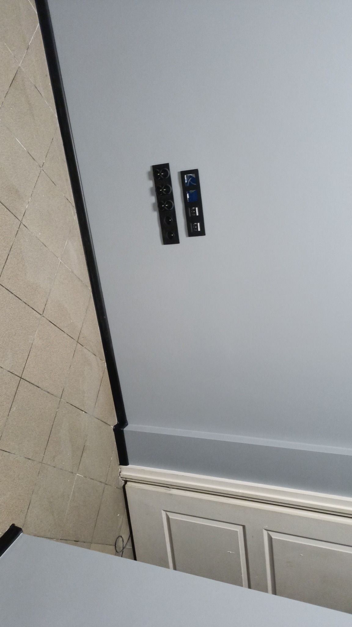 Świeżo zamontowane, czarne gniazdka elektryczne i porty USB na szarej ścianie nad białą listwą przypodłogową i beżową podłogą z płytek.