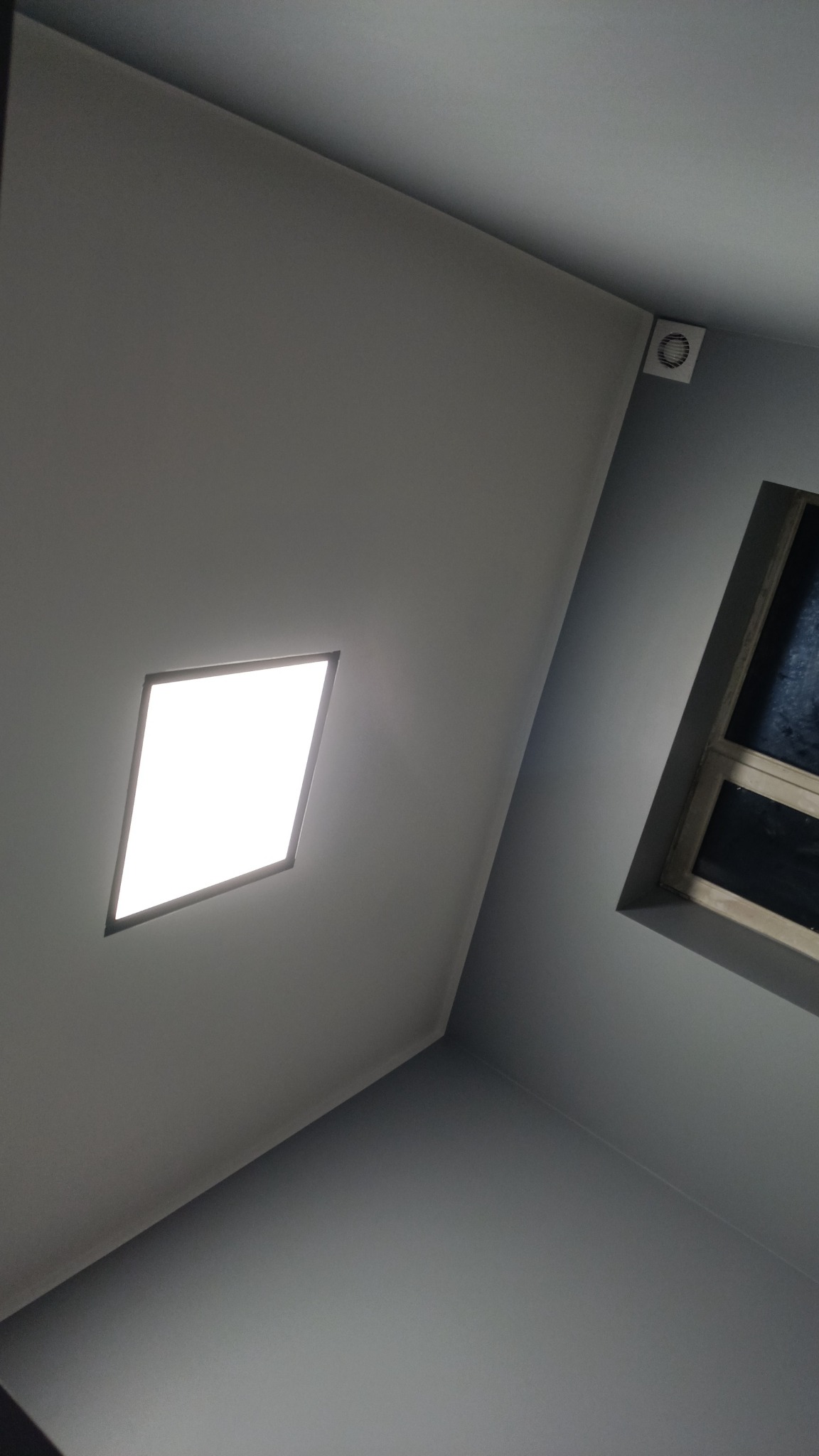 Szare wnętrze z widokiem na sufit z kwadratową lampą LED i oknem w ramie, widoczny także wentylator sufitowy.