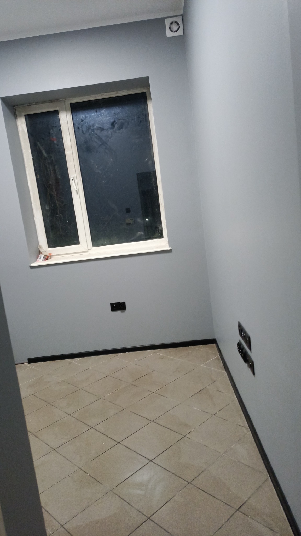 Pokój po remoncie z szarymi ścianami, beżowymi płytkami podłogowymi, białym oknem i czarnymi listwami przypodłogowymi oraz gniazdkami elektrycznymi.