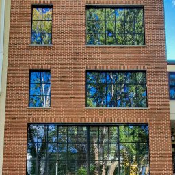 ALU-HOUSE Brzycki - Elewacja trzypiętrowego budynku z czerwonej cegły z dużymi, czarnymi oknami, w których odbija się zieleń drzew i błękitne niebo.