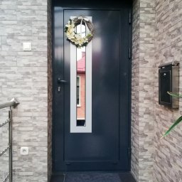 ALU-HOUSE Brzycki - Grafitowe drzwi wejściowe z podłużnym przeszkleniem, udekorowane wiankiem z jasnych kwiatów, na tle ściany wyłożonej szarą cegłą imitującą kamień.