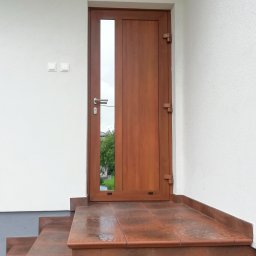 ALU-HOUSE Brzycki - Drewniane drzwi wejściowe z szybą, zamontowane na zewnątrz budynku z białymi ścianami i brązowymi schodami z płytek.
