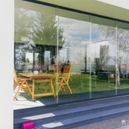 Szklana ściana przesuwna oddzielająca taras z drewnianym stołem i krzesłami od zielonego ogrodu z widocznym grillem i drzewami.