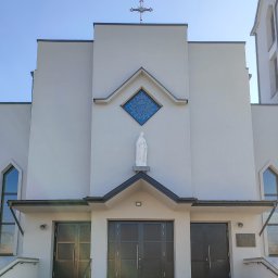 ALU-HOUSE Brzycki - Fasada kościoła z trzema parami ciemnych drzwi zewnętrznych, nad środkowymi drzwiami statua Matki Boskiej i witraż w kształcie rombu, na szczycie krzyż.