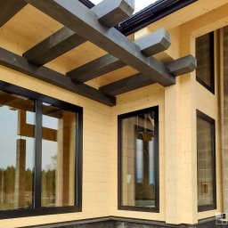 ALU-HOUSE Brzycki - Nowoczesny dom z jasnego drewna z czarnymi ramami okiennymi, widoczne także ciemne drewniane belki stropowe i fragment murowanego komina.