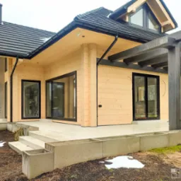 Nowoczesny dom z jasnego drewna z czarną dachówką, czarnymi ramami okien i przesuwnymi drzwiami, oraz ciemną drewnianą pergolą