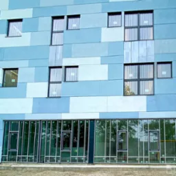 Fasada budynku z oknami aluminiowymi o zróżnicowanych rozmiarach i układach, na tle niebieskich paneli elewacyjnych w różnych odcieniach.