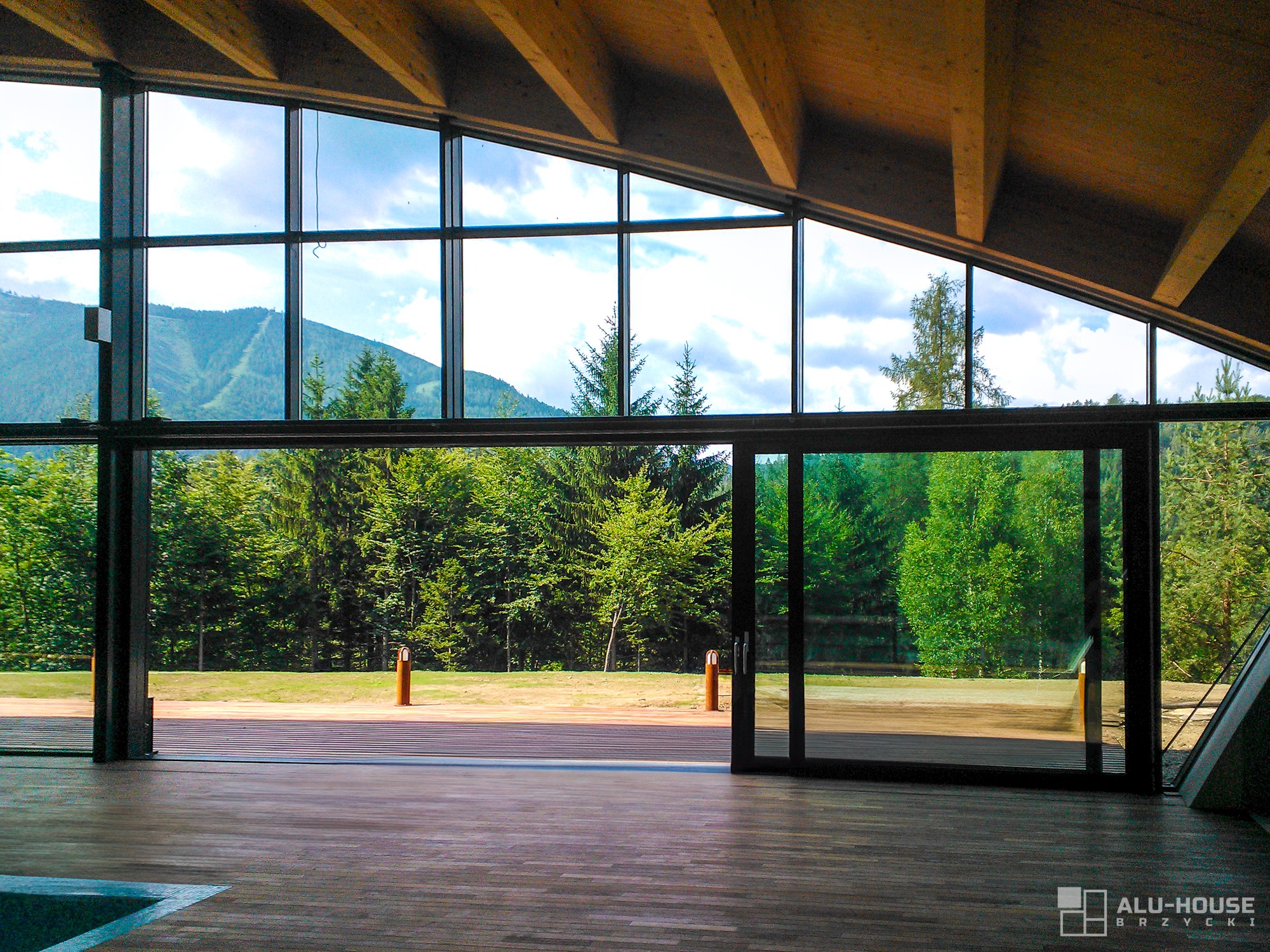 Otwarta, panoramiczna aluminiowa konstrukcja przesuwna z widokiem na las i góry, drewniana podłoga wewnątrz, nowoczesny design.