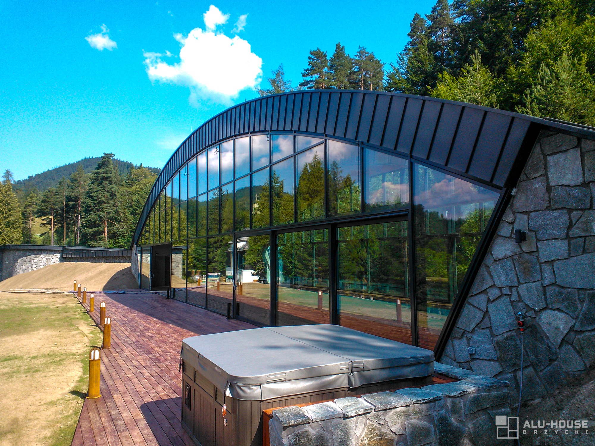 Nowoczesny budynek z zakrzywioną fasadą z szklanych paneli, otoczony zielenią i drewnianym tarasem z jacuzzi, widok z zewnątrz w słoneczny dzień.