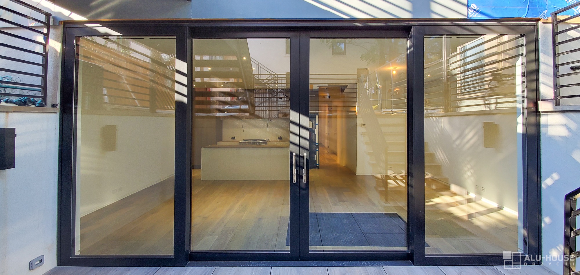 Nowoczesne, czarne drzwi przesuwne z dużymi szybami, widok na wnętrze domu z drewnianą podłogą, schodami i kuchnią w tle, oświetlone promieniami słońca padającymi przez żaluzje.