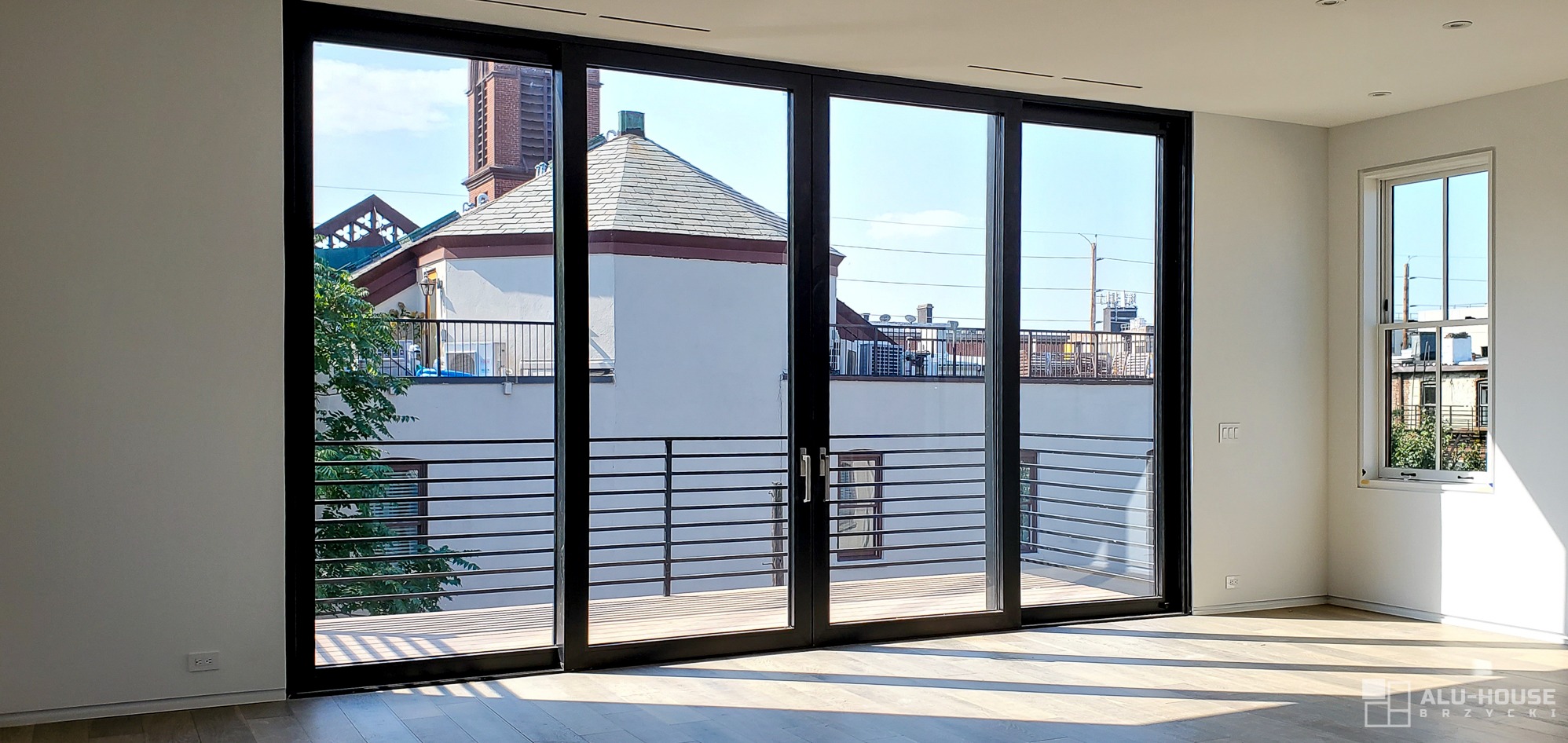 Wnętrze z dużymi, przesuwnymi, czarnymi drzwiami tarasowymi z widokiem na budynek z ceglaną wieżą i balkonem, oraz dodatkowym oknem po prawej stronie.