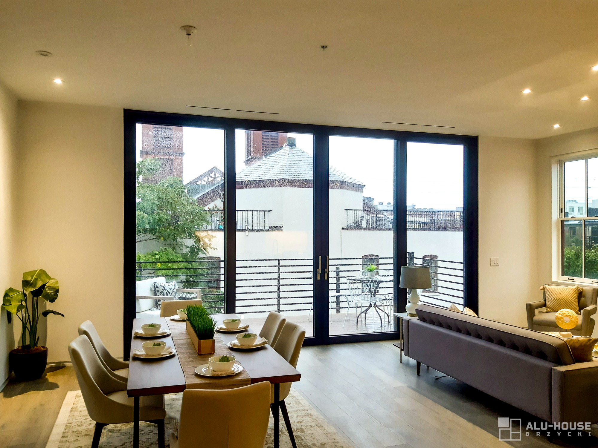 Jasne wnętrze z dużymi przesuwnymi oknami z czarnymi ramami, widok na balkon i zabudowania miejskie, stół jadalny z nakryciem, szara sofa i logo firmy w prawym dolnym rogu.