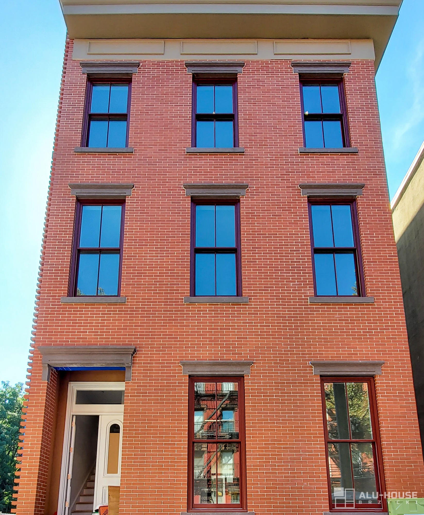 Fasada budynku z czerwonej cegły z nowymi oknami w bordowych ramach, widoczny fragment wejścia z białą stolarką.