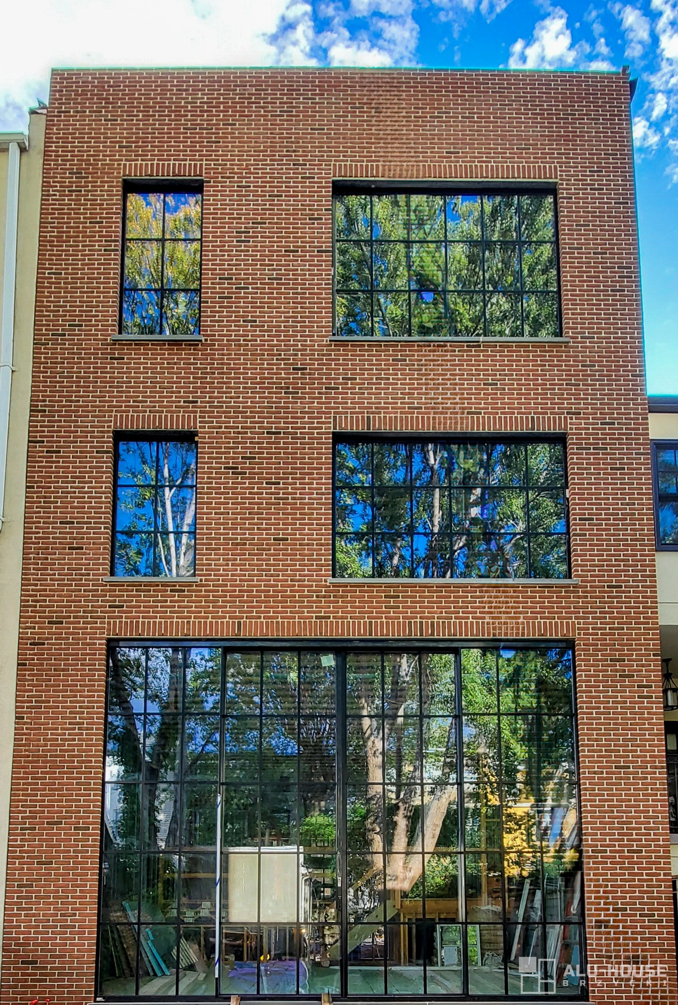 Elewacja trzypiętrowego budynku z czerwonej cegły z dużymi, czarnymi oknami, w których odbija się zieleń drzew i błękitne niebo.