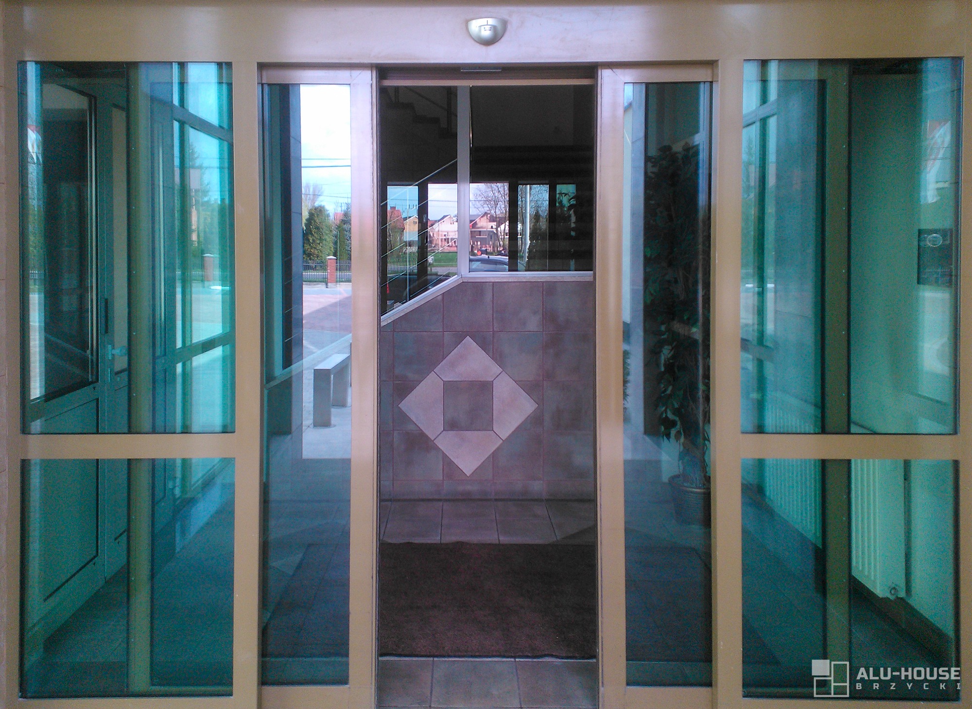 Szklane drzwi przesuwne z aluminiową ramą, widok na wnętrze z kafelkową ścianą i wzorem rombu, oraz zewnętrzny krajobraz z domami i zielenią.