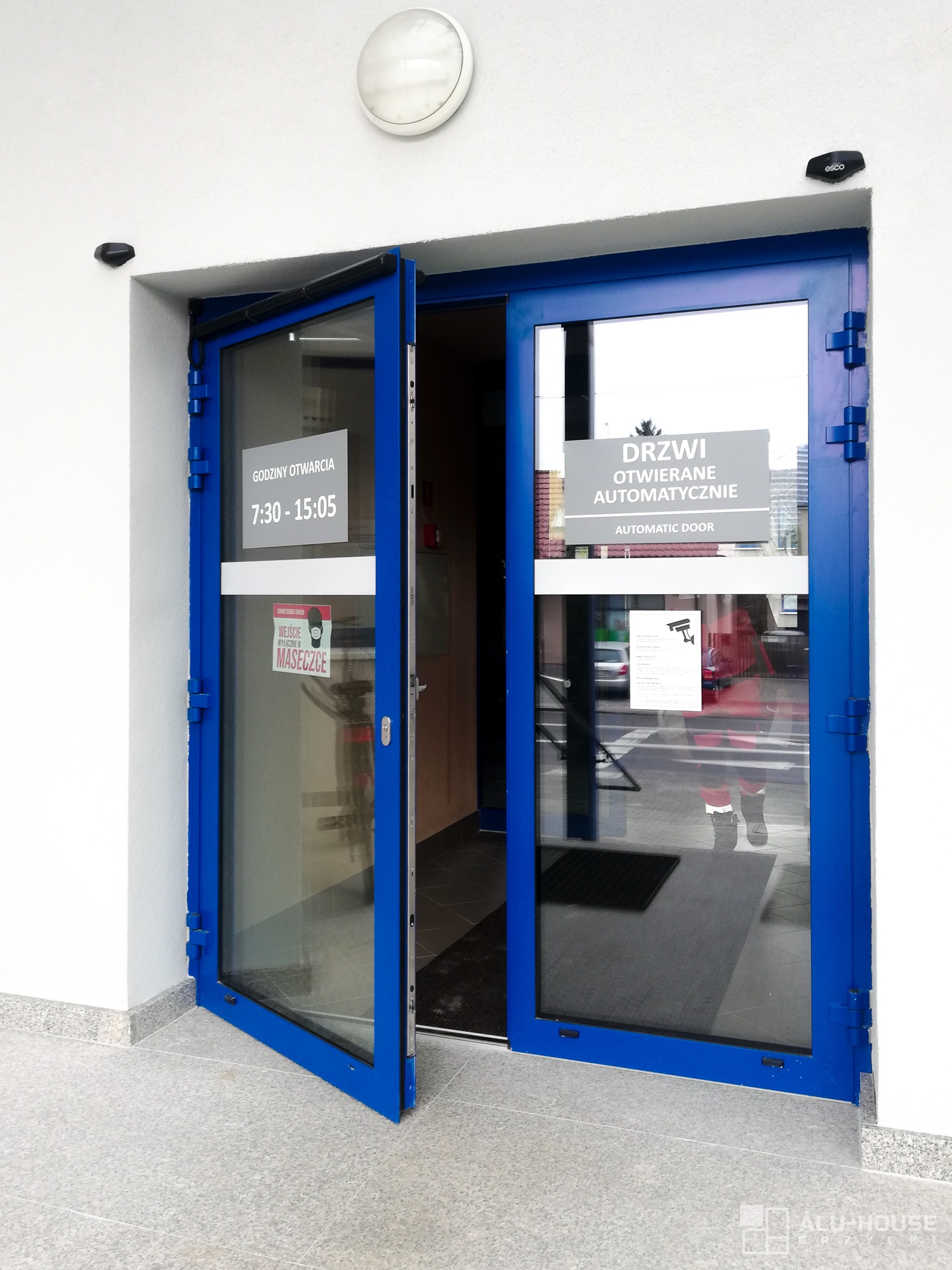 Automatyczne drzwi wejściowe z niebieską ramą, częściowo otwarte, z widocznym napisem 'Godziny otwarcia' i informacją o obowiązku noszenia maseczki, zamontowane w Ropczycach.