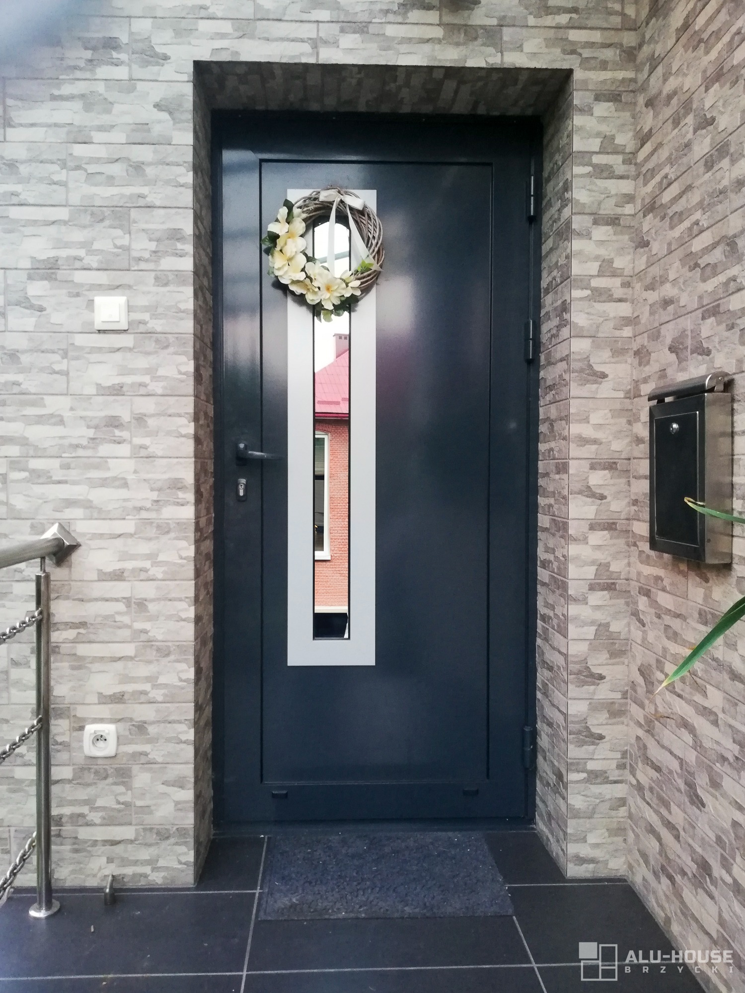 Grafitowe drzwi wejściowe z podłużnym przeszkleniem, udekorowane wiankiem z jasnych kwiatów, na tle ściany wyłożonej szarą cegłą imitującą kamień.
