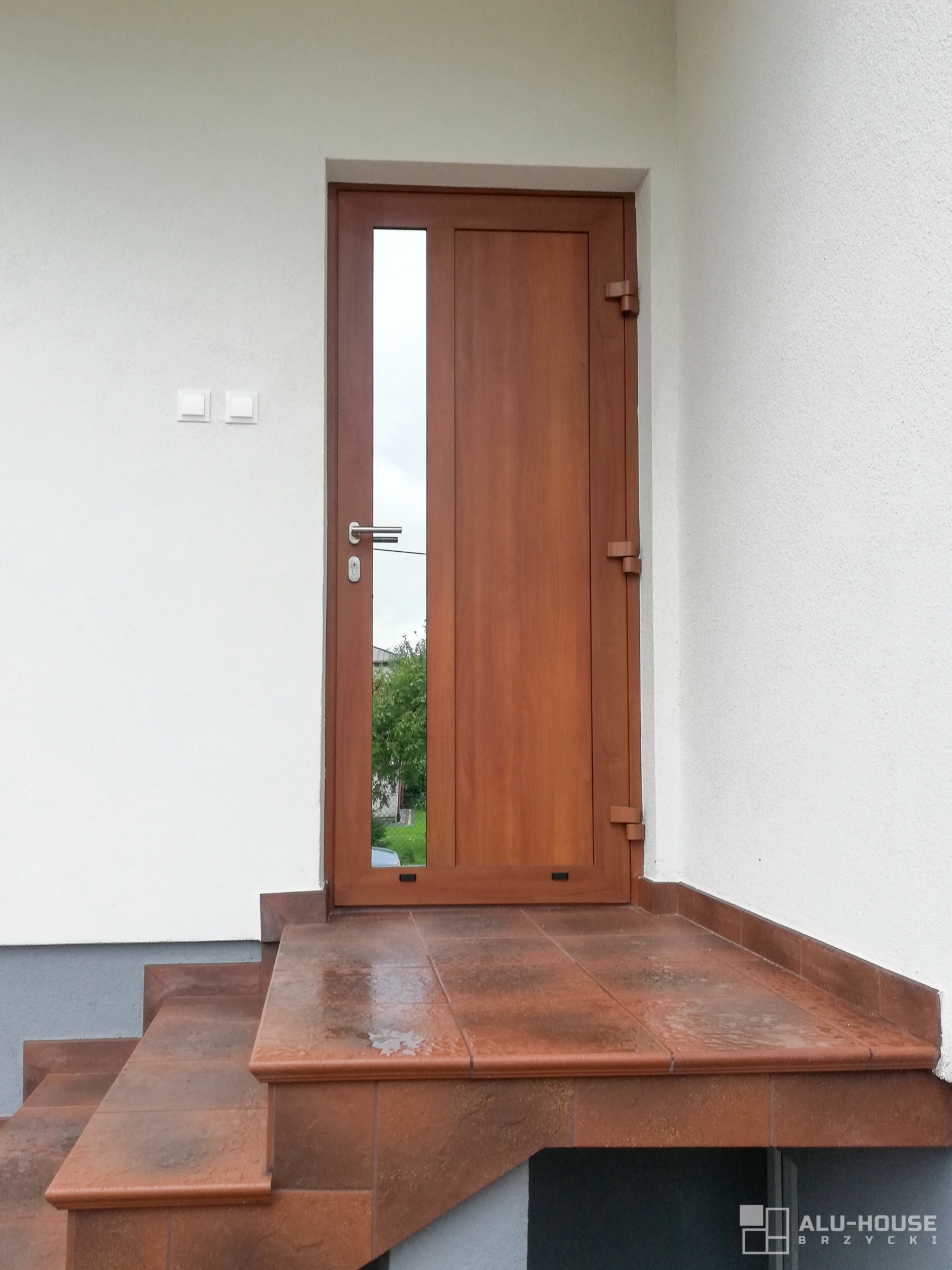 Drewniane drzwi wejściowe z szybą, zamontowane na zewnątrz budynku z białymi ścianami i brązowymi schodami z płytek.