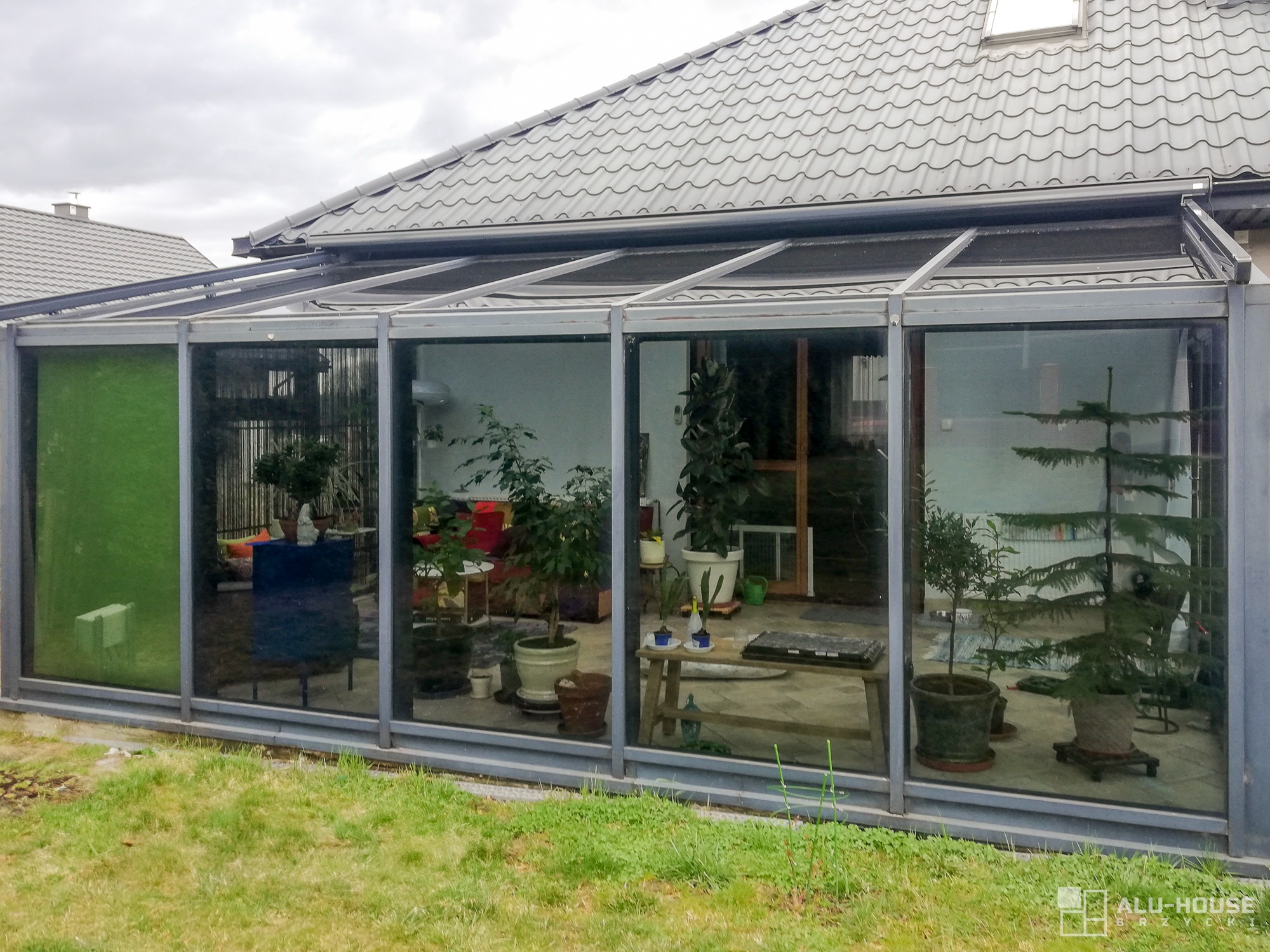 Szklana zabudowa tarasu z metalową konstrukcją, widok z zewnątrz, wewnątrz rośliny doniczkowe i meble ogrodowe, na zewnątrz trawnik.