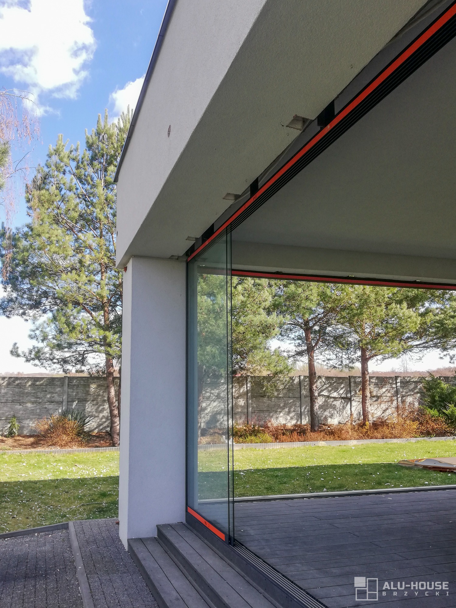 Nowoczesne, bezramowe przeszklenie tarasowe z widokiem na ogród, z elementami wykończeniowymi w kolorze pomarańczowym, widoczne schody z desek kompozytowych.