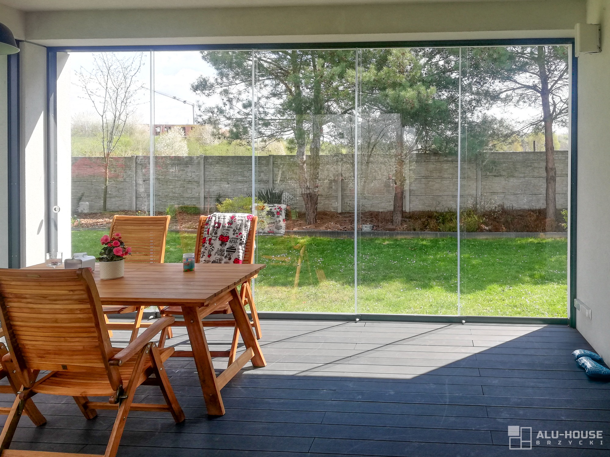Zadaszony taras z drewnianym stołem i krzesłami, oddzielony od ogrodu szklanymi, przesuwnymi panelami bezramowymi, zapewniającymi panoramiczny widok na zieleń.