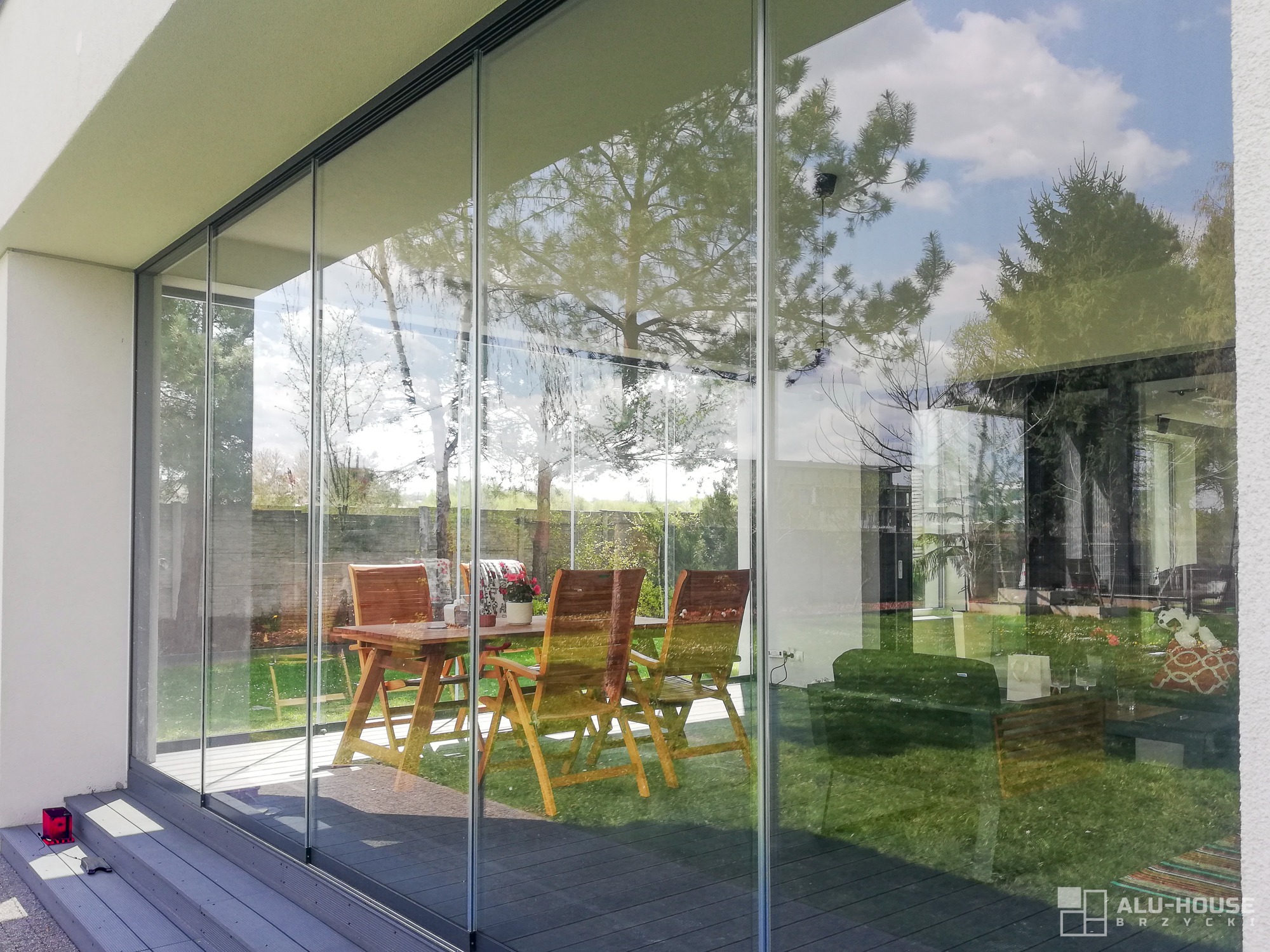 Szklane drzwi przesuwne tarasowe z widokiem na drewniany stół i krzesła ogrodowe na trawniku, refleksja drzew i nieba w szkle, nowoczesny design.